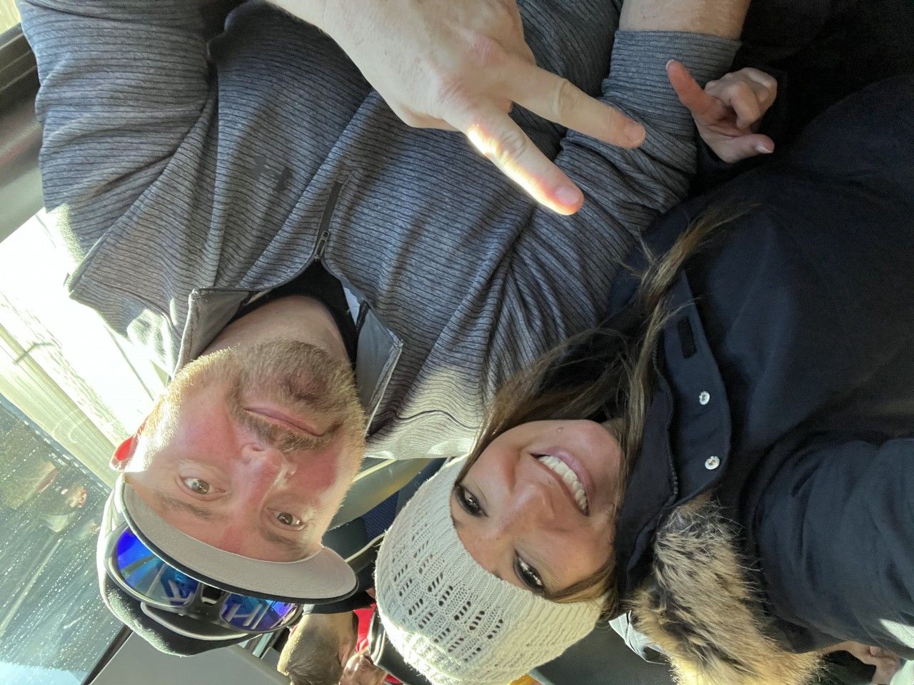 Two smiling people taking a selfie; man makes a peace sign. They are indoors by a window.