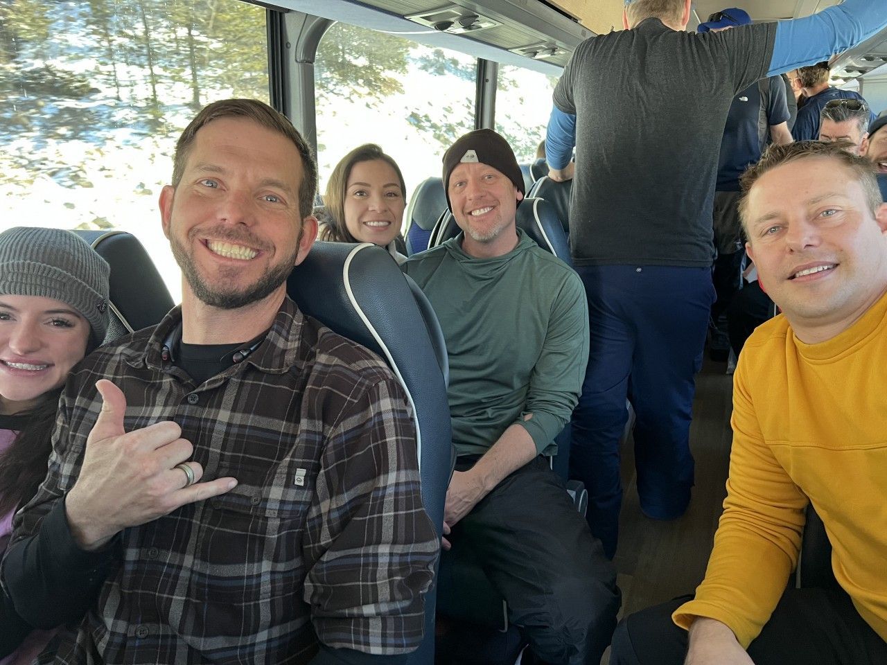 People smiling inside a bus. Man in plaid shirt gives hang loose sign. Snowy landscape seen through windows.