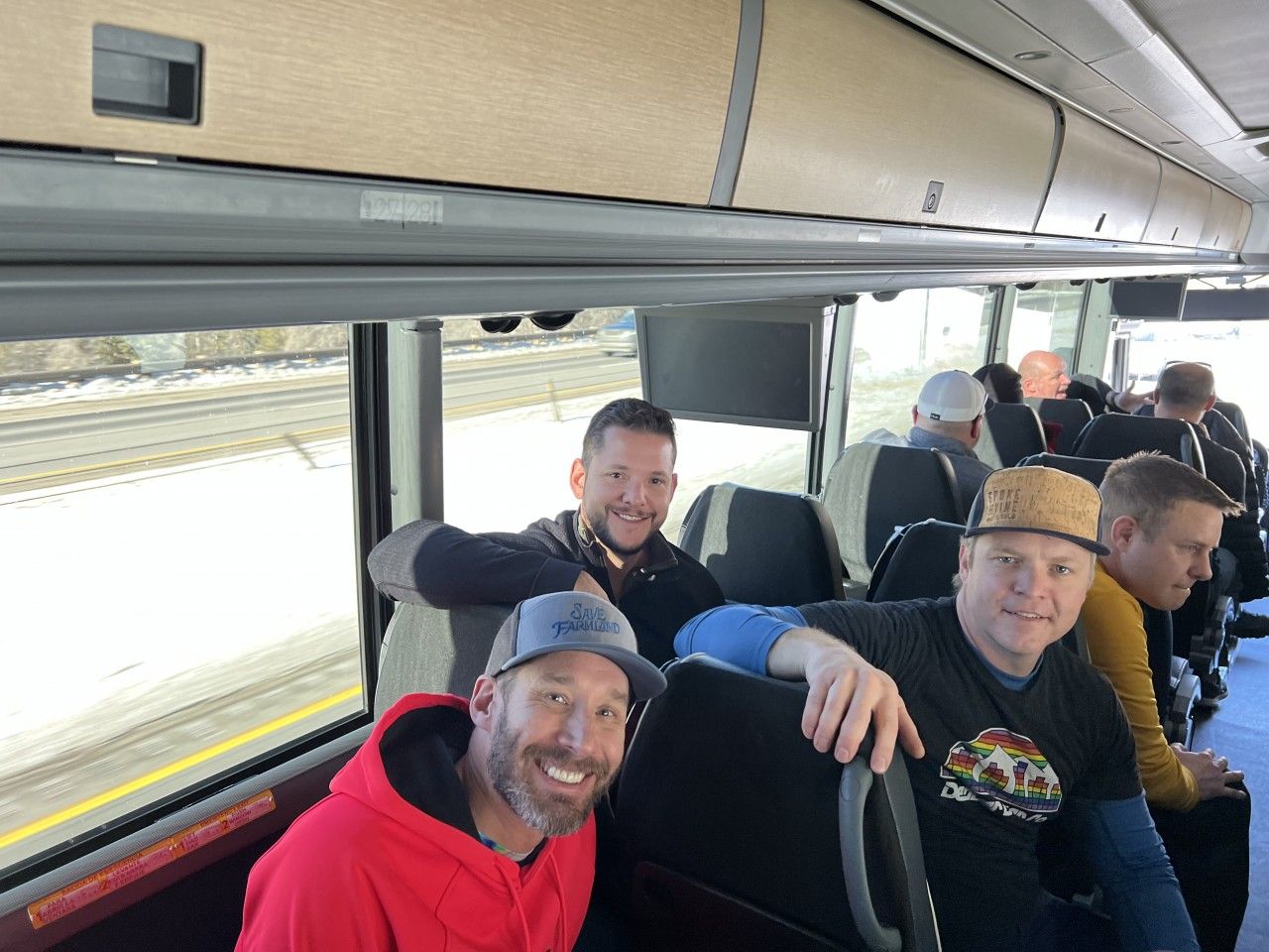 Three men smiling on a bus with other passengers. One in red hoodie, another with a beard, and another in a t-shirt and hat.