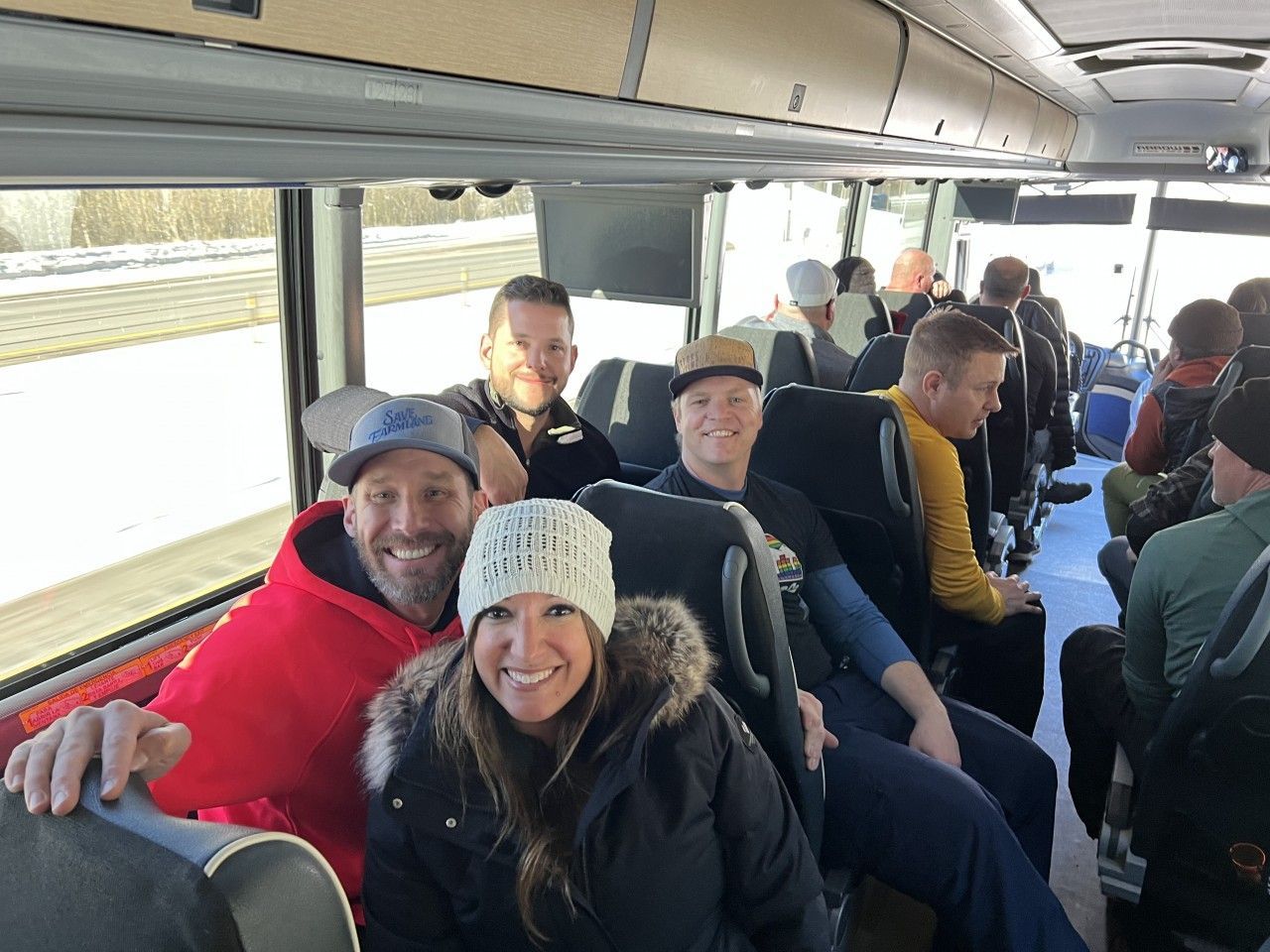 People sitting on a bus; some smiling. Bright day, snowy scene visible through the windows.