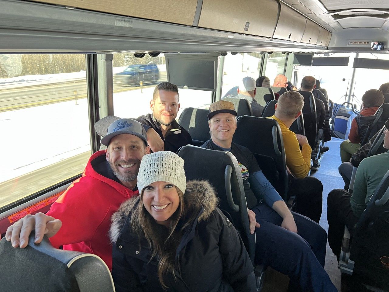 People on a bus, smiling. One in a red jacket, seated near a person in a white hat and black coat.