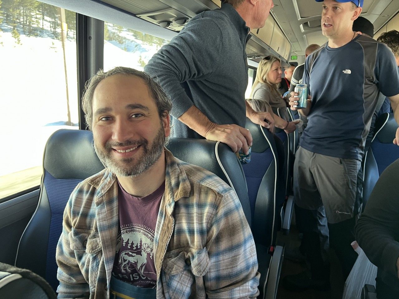 Man smiles on a bus, wearing plaid shirt. Passengers stand in the background.