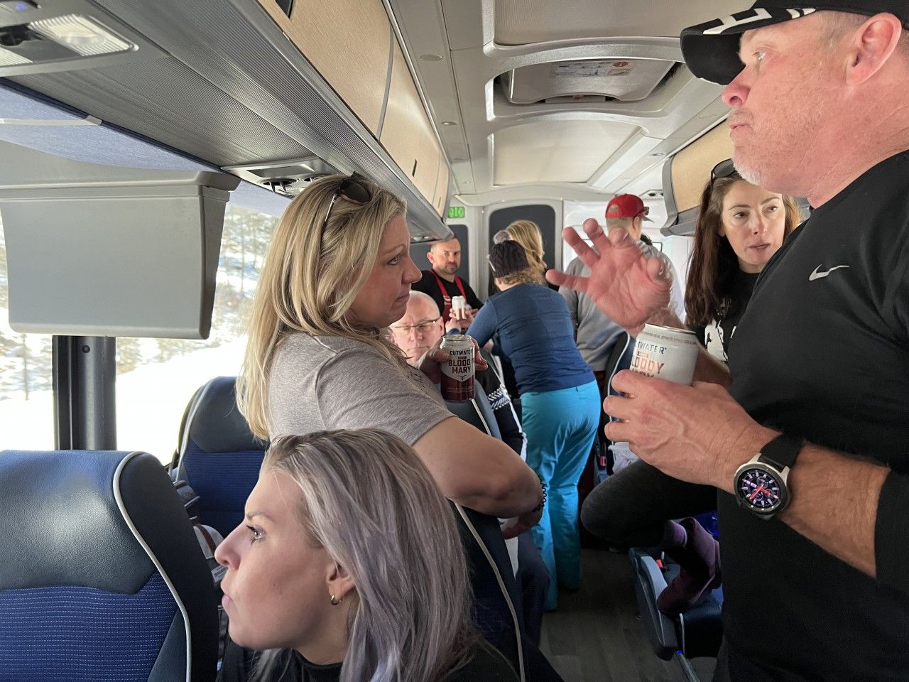 People on a bus, talking. Interior shot. Some are holding cups, one is gesturing.