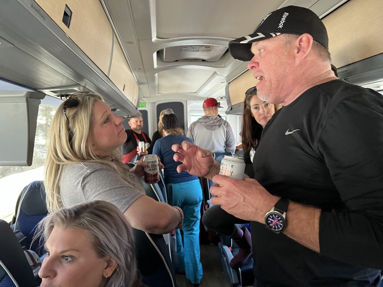People talking on a bus. A man gestures, holding a cup. Others stand and sit, some holding beverages.
