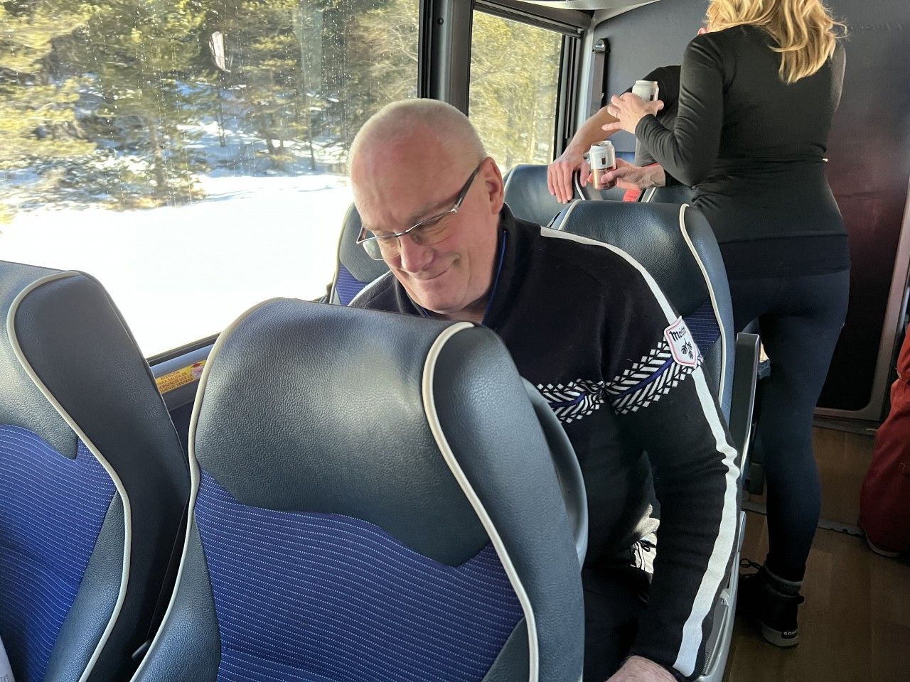 Man in glasses on a bus, smiling. Another person adjusts their hair. Blue and black seats. Snow outside window.