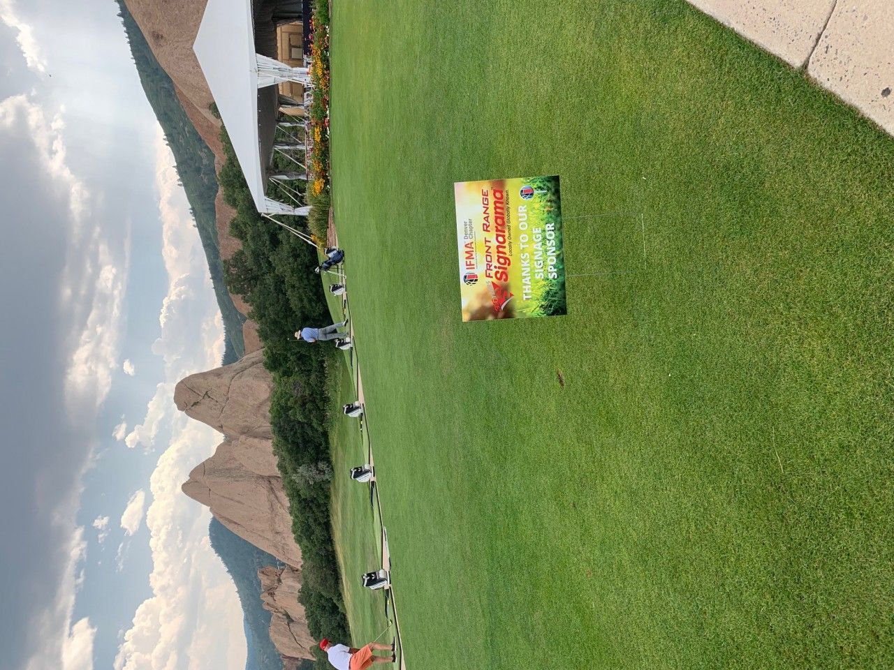 Sign on green grass with mountains and blue sky in the background.