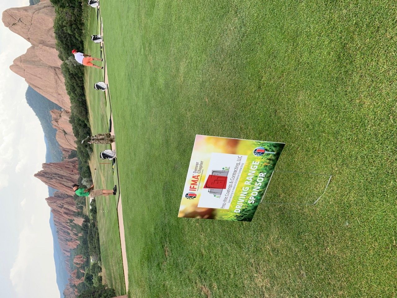 Green golf course with sign, mountains, and people.