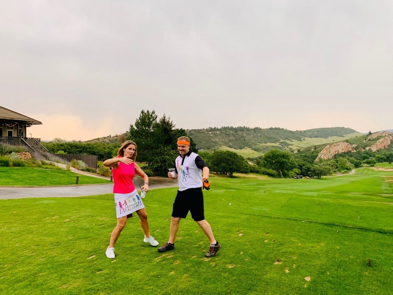 Woman in pink and man in white t-shirt on a golf course, both posing, possibly celebrating, with hills in the background.