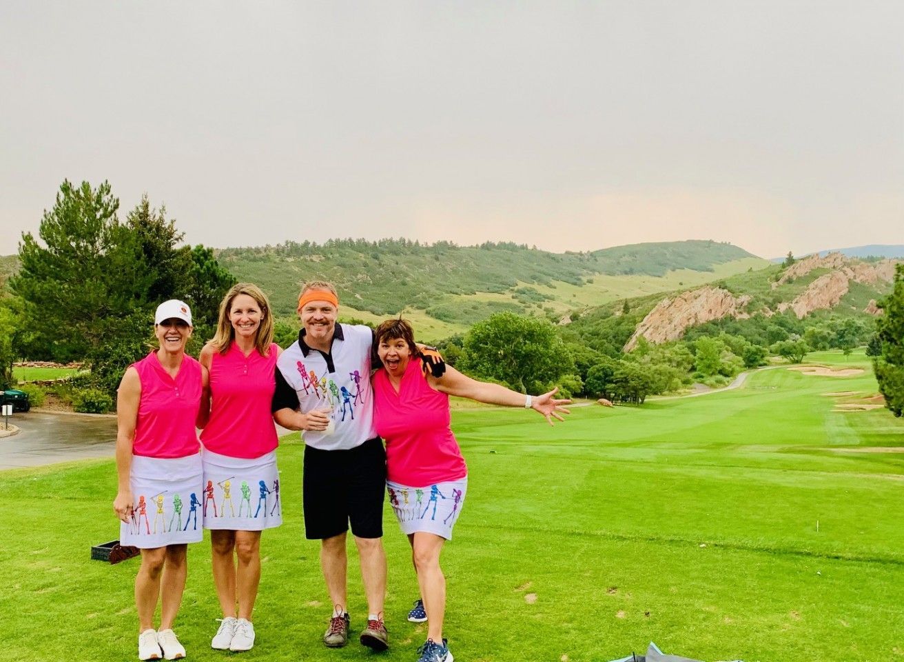 Group of four people on a golf course, three wearing matching pink shirts and white skirts, one man holding a trophy.