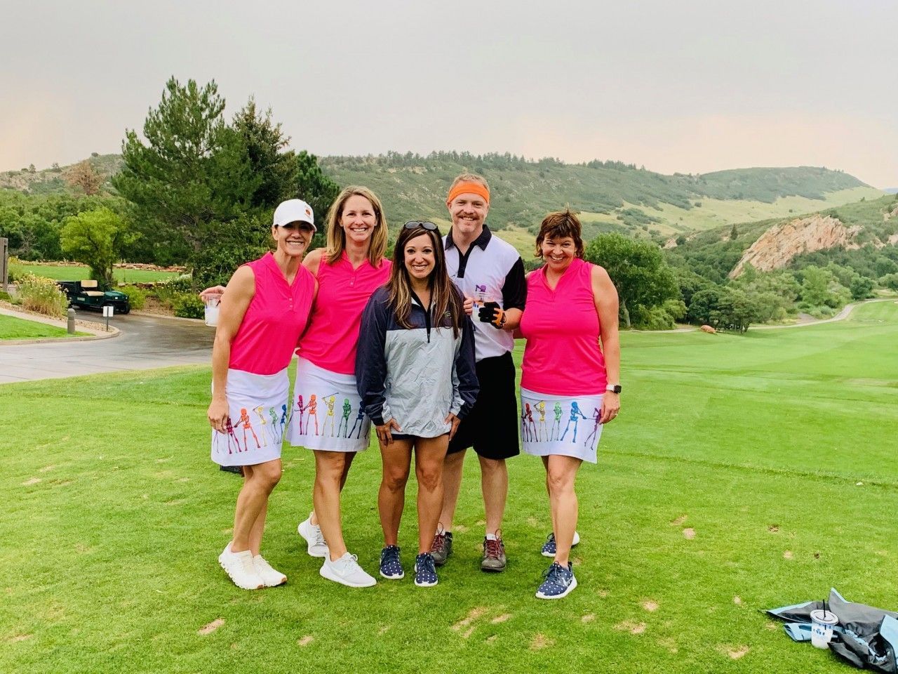 Group of golfers in pink shirts and patterned skirts stand on a green golf course, smiling.