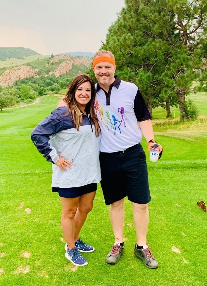 A couple smiling on a golf course. Man in black shorts and a colorful shirt, woman in a jacket and shorts.