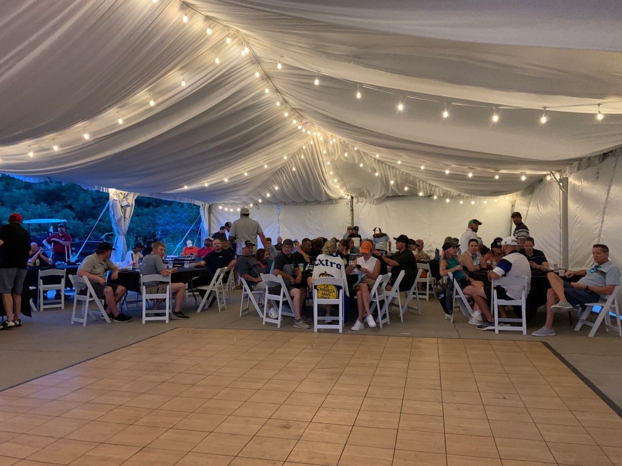 People gather under a tent with string lights and a dance floor. Tables and chairs are set up for dining.