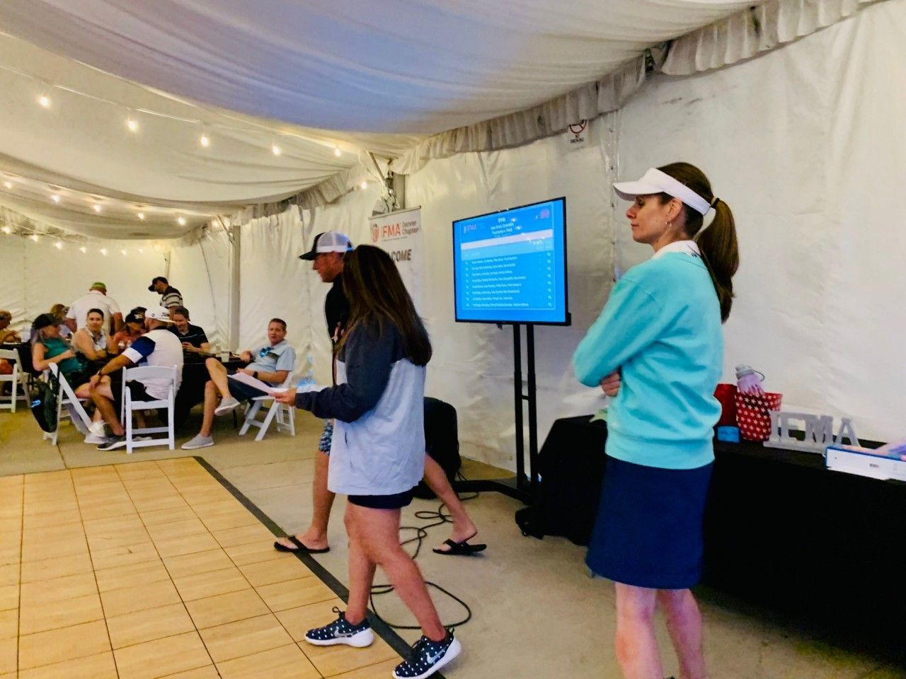 People in tent, presenting to audience. Woman in blue, man with hat, presentation screen visible.