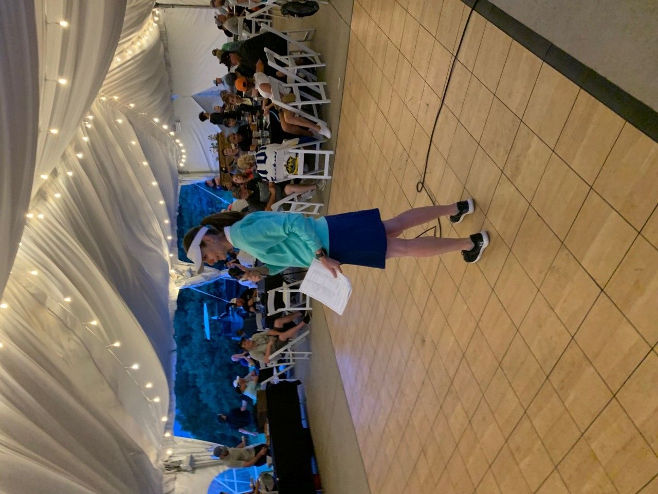 Woman speaking at event; microphone, paper in hand; inside tent, seated audience, string lights, wood floor.