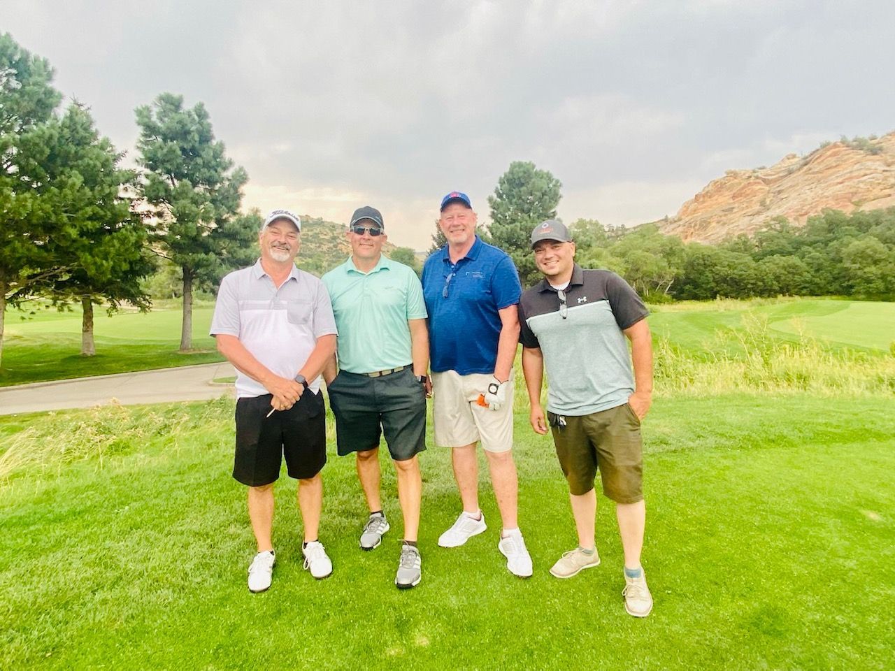 Four men smiling on a golf course. They are wearing golf attire, standing on green grass with trees and a cloudy sky.