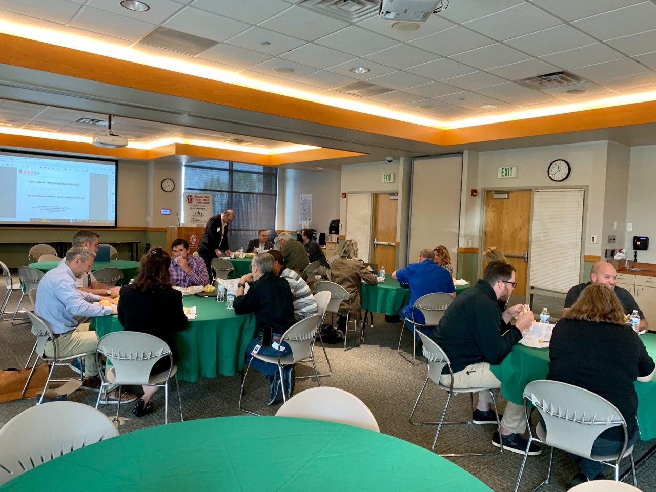 Group of people in a meeting room, sitting at green round tables. A man stands, possibly presenting.
