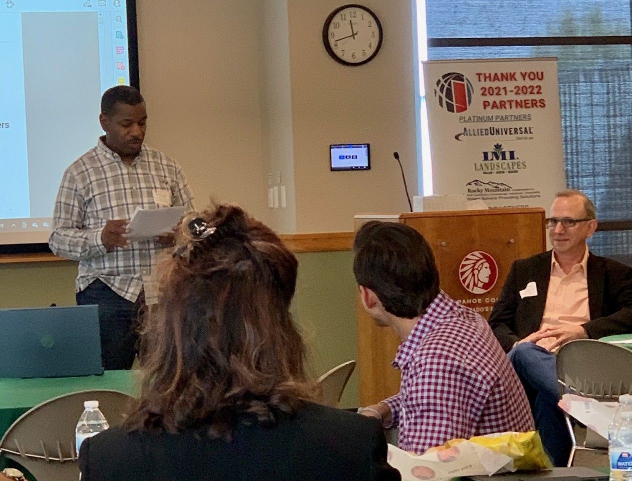 Man presenting at a conference, others seated in the audience.