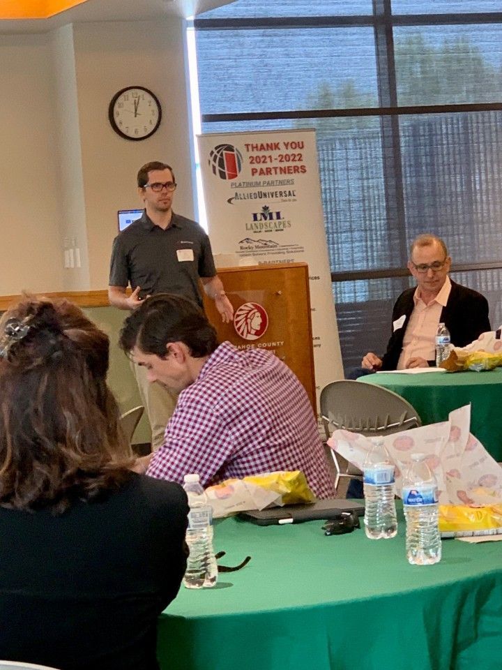 Man speaking at a podium, two others at a table with green tablecloth. A banner behind the speaker.