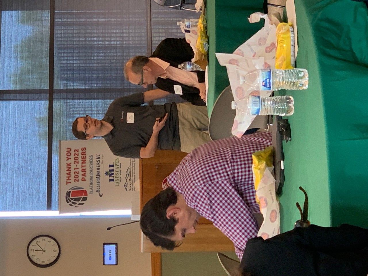 People at a table in a room. One man gestures, others look down at the table. Food and water are present.