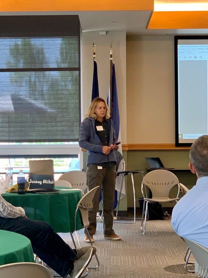 Woman speaking at an event, wearing a jacket and pants, flags in background, laptop, and seated audience.