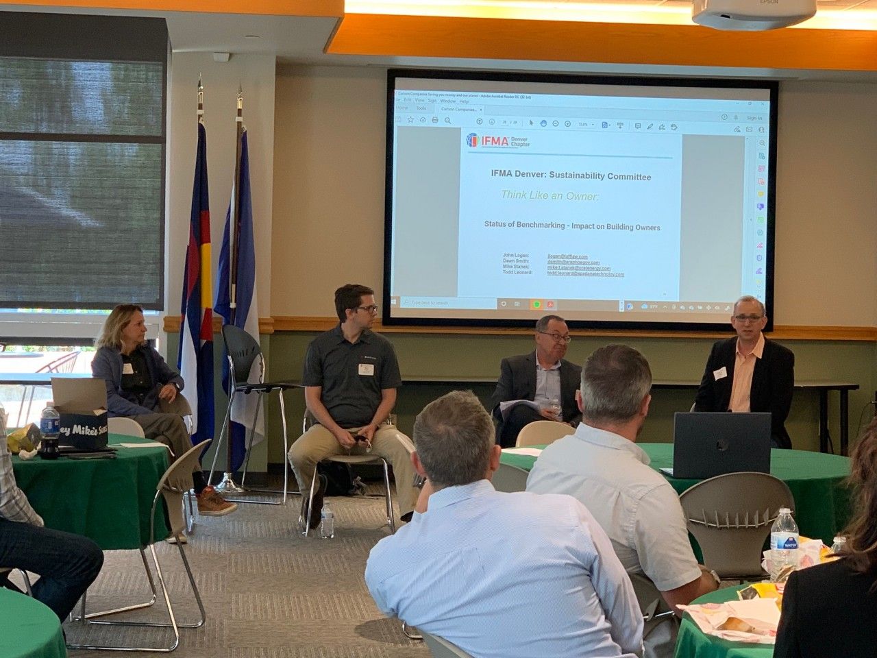 People seated at tables in a conference room, with presenters at the front; a screen displays a document.