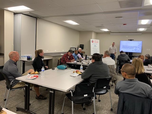 People at a meeting, seated around tables. A man presents to the group, a screen visible behind him.