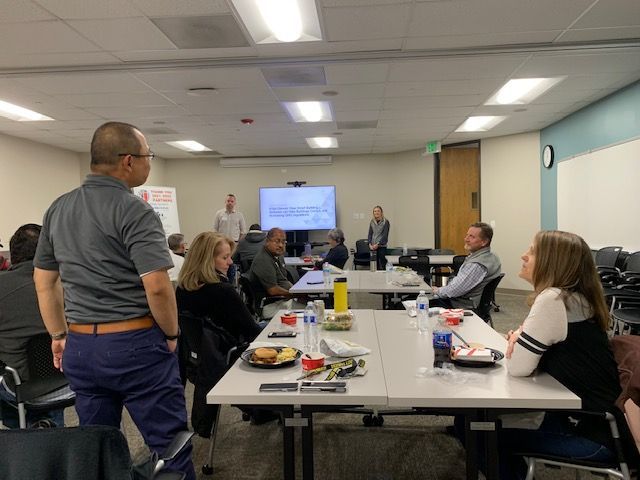 People in a meeting room, seated at tables and facing a presentation on a screen.