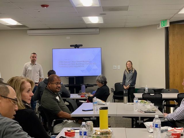 People in a meeting room, two at the front. A presentation on the screen. Attendees at tables.