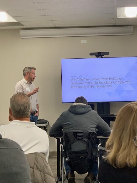 Man giving a presentation. Audience seated, one person in a wheelchair. A screen displays text.