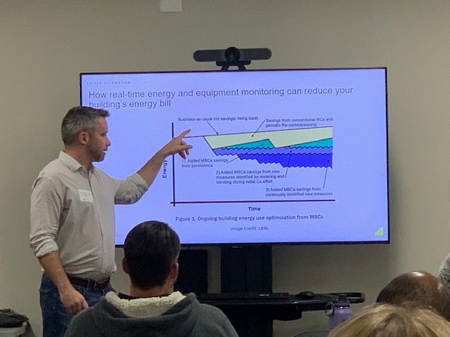 Man presenting a graph on a screen, pointing to it, in front of an audience.