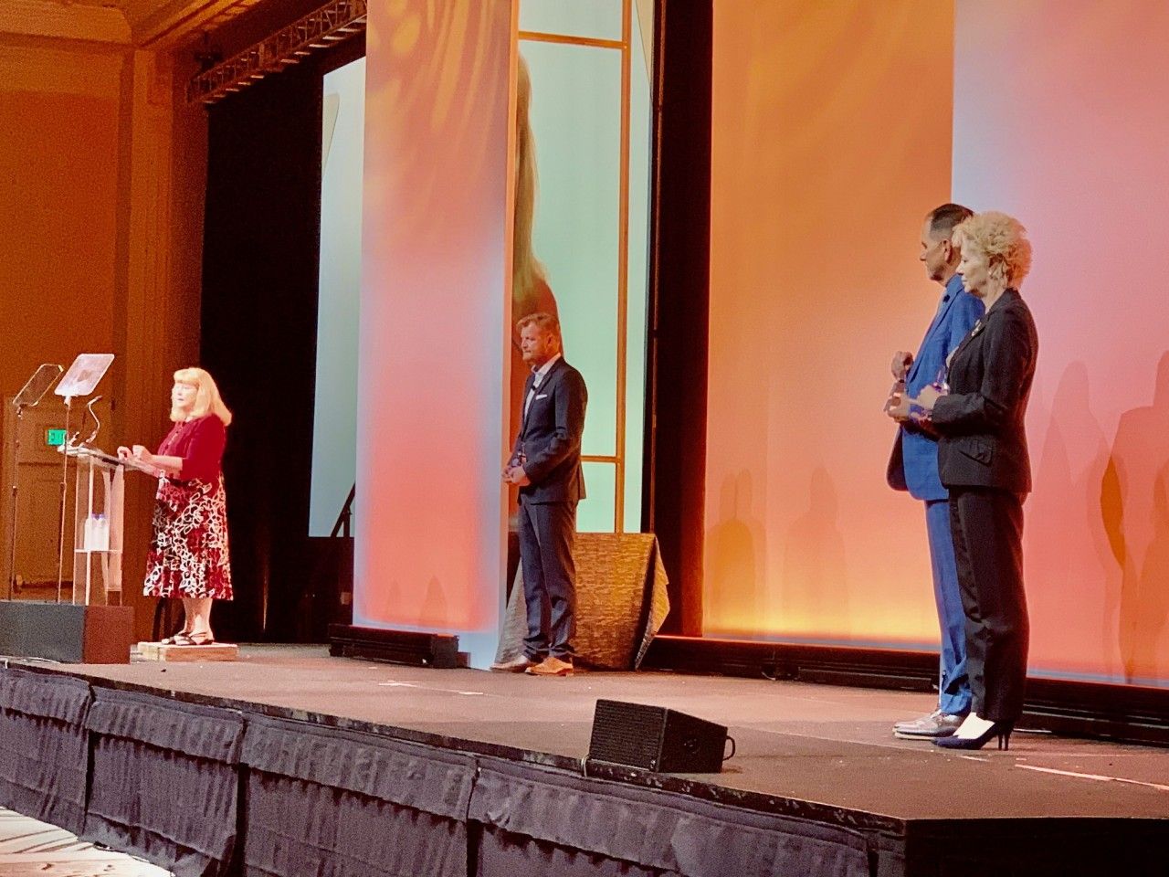 Woman speaking at a podium; two men and a woman stand behind her on a stage with a backdrop.