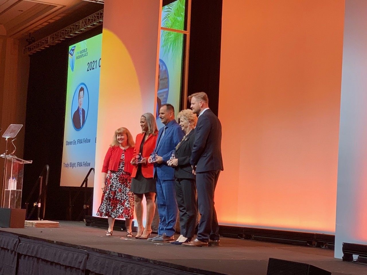 People on stage at an awards ceremony; two hold trophies. Lit stage with orange backdrop.