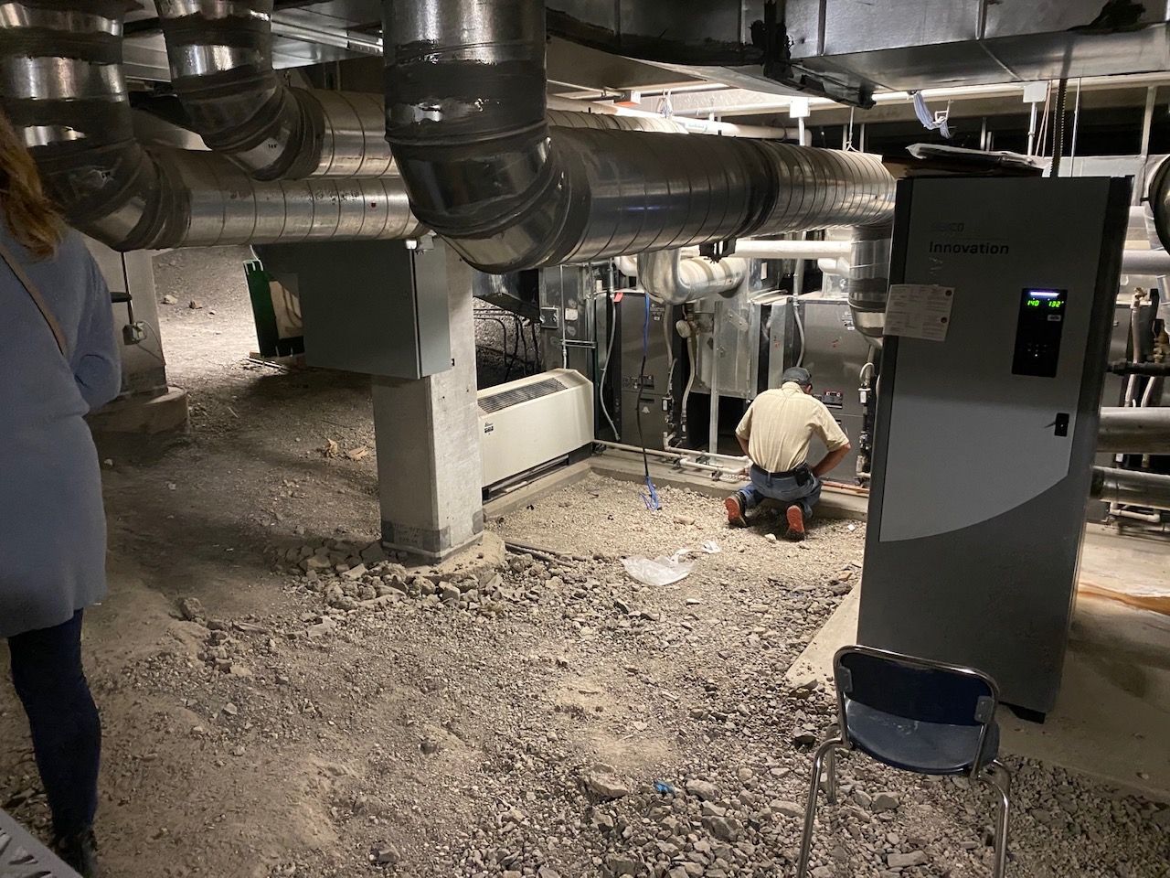 Person working on machinery in a basement with pipes and dusty ground.