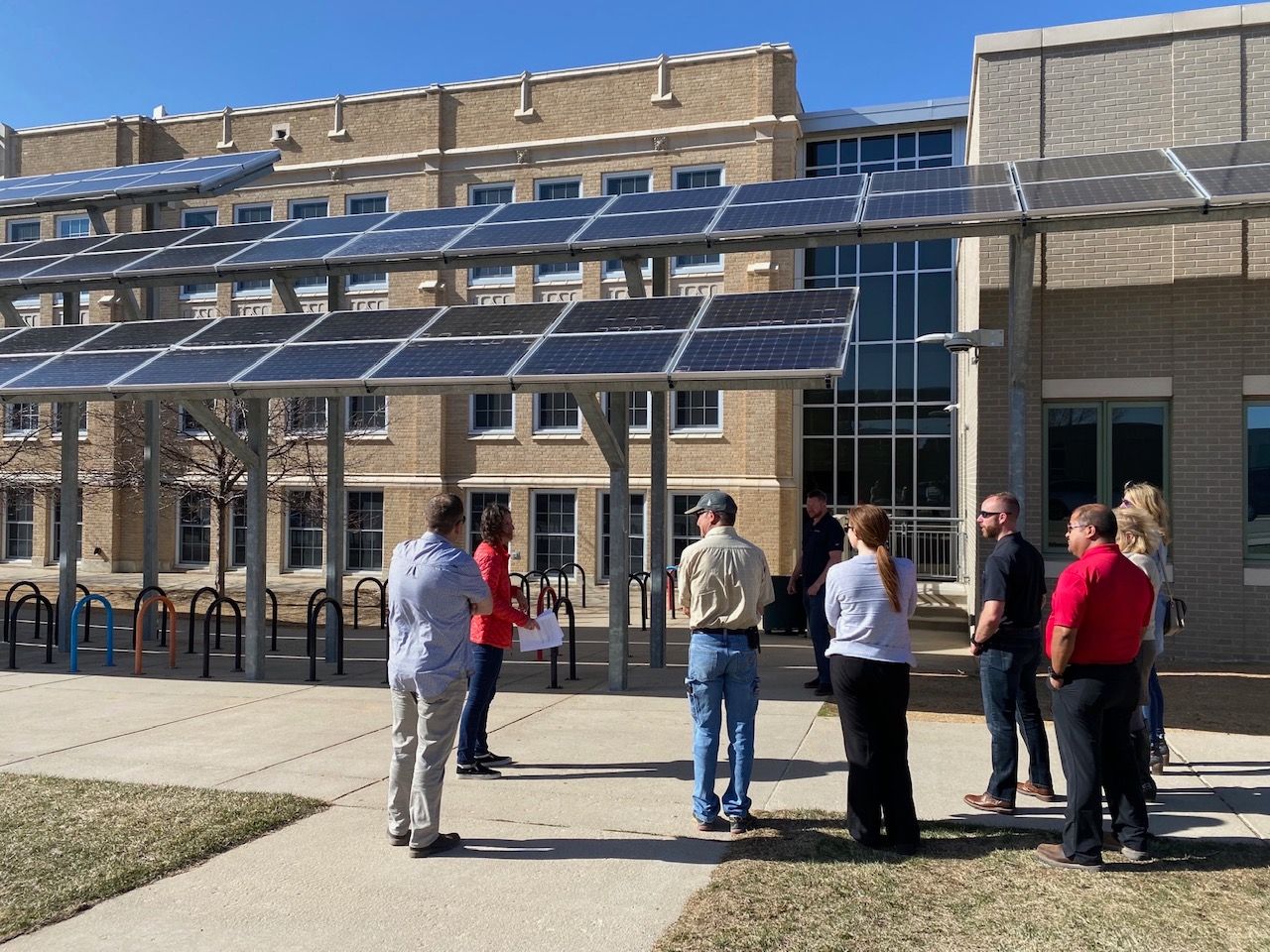 People gather under solar panels outside a building with brick facade.