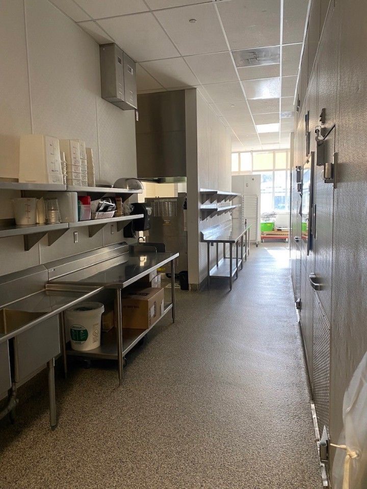 Commercial kitchen with stainless steel countertops, sinks, and shelves. Long hallway with refrigerators on the right.