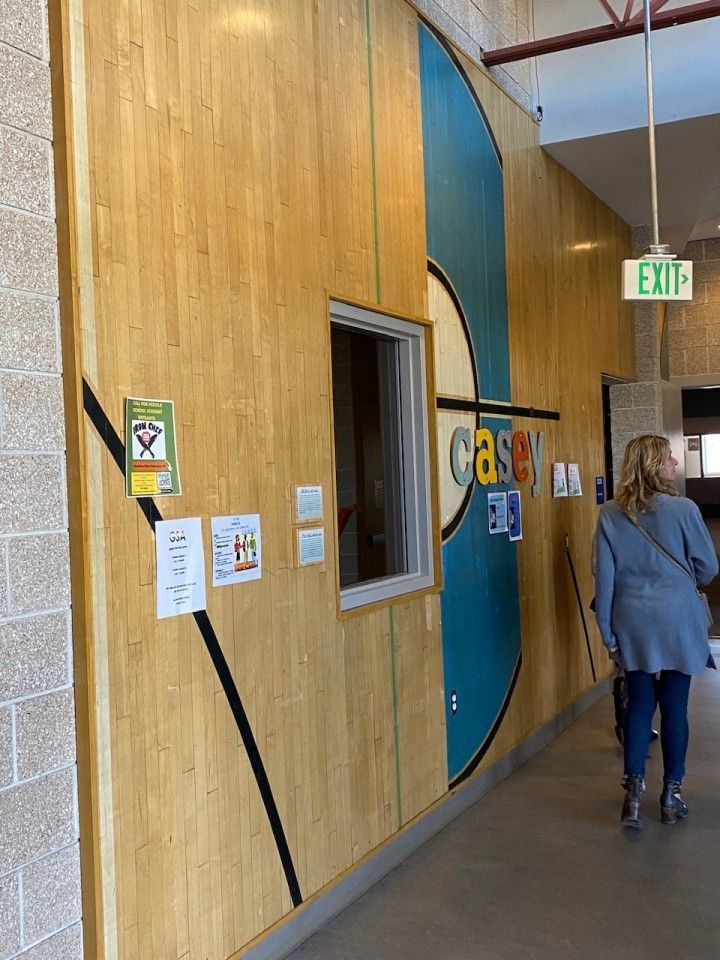 Wood-paneled wall with a teal graphic and small window. A woman walks down the hallway past an exit sign.