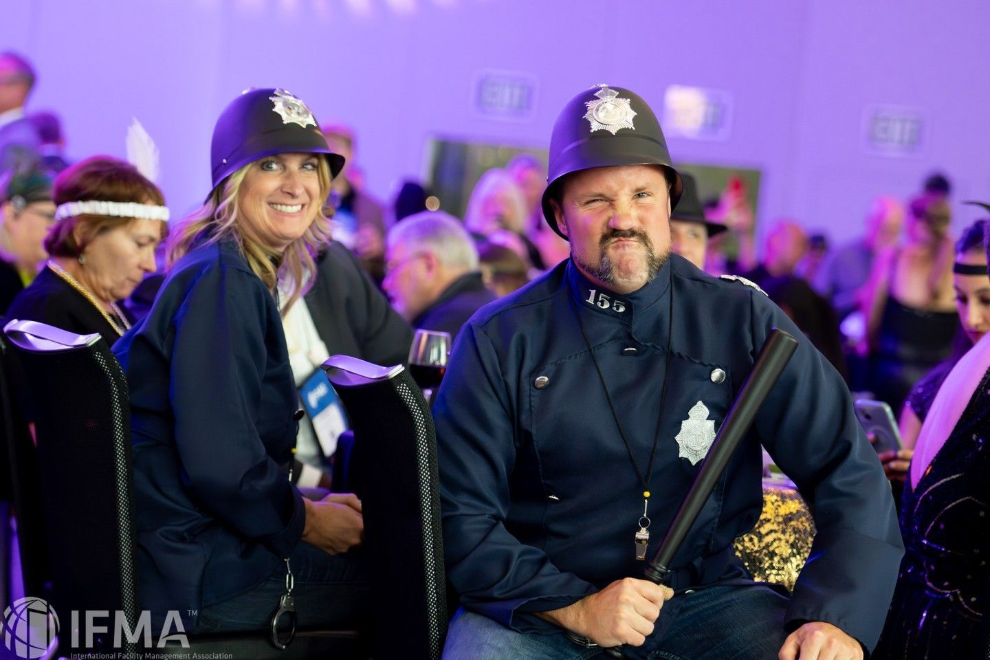 Two people in police officer costumes at an event, one holding a baton, both smiling, seated.