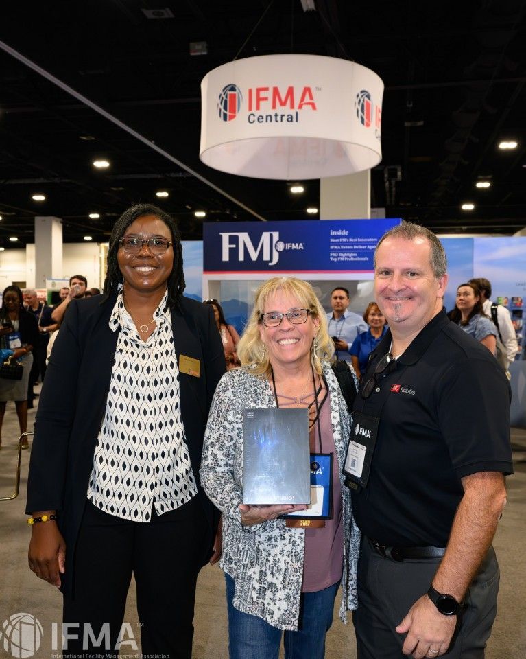 Three people pose with a trophy at the IFMA Central booth at a conference.