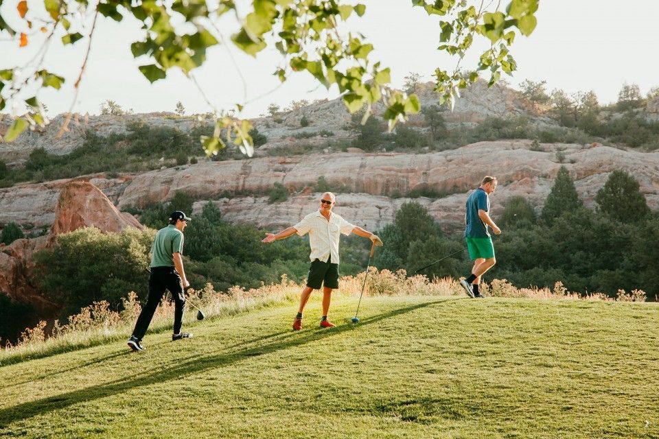 Three people golfing on a green hill with red rock formations and trees in the background.