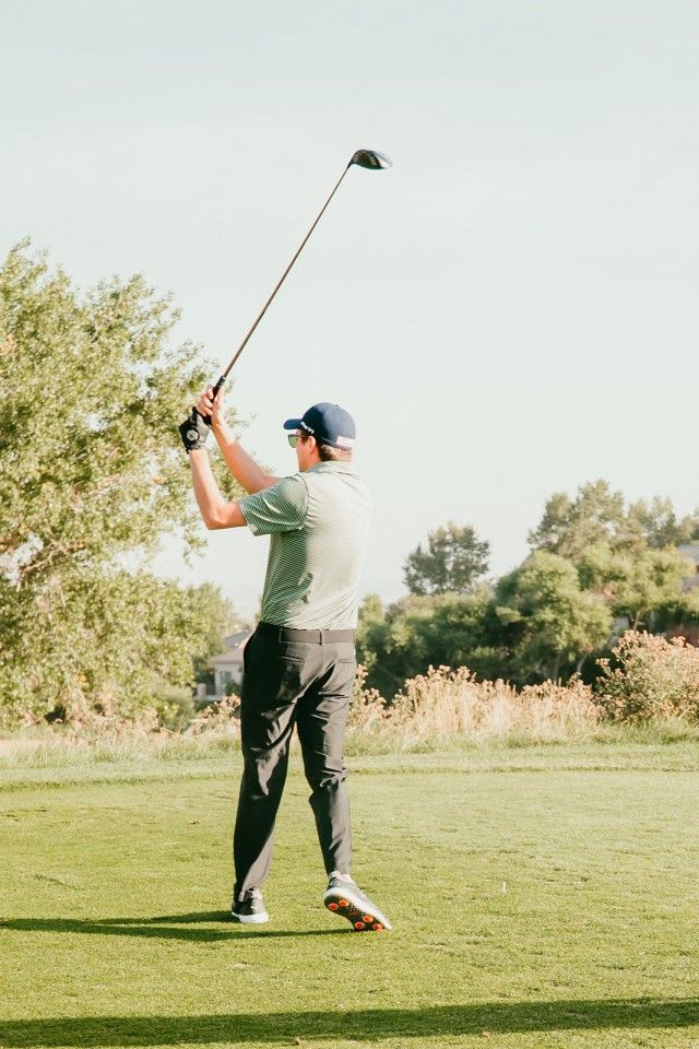 Golfer swinging a club on a green lawn, wearing a cap, shirt, and dark pants; sunny outdoor setting.