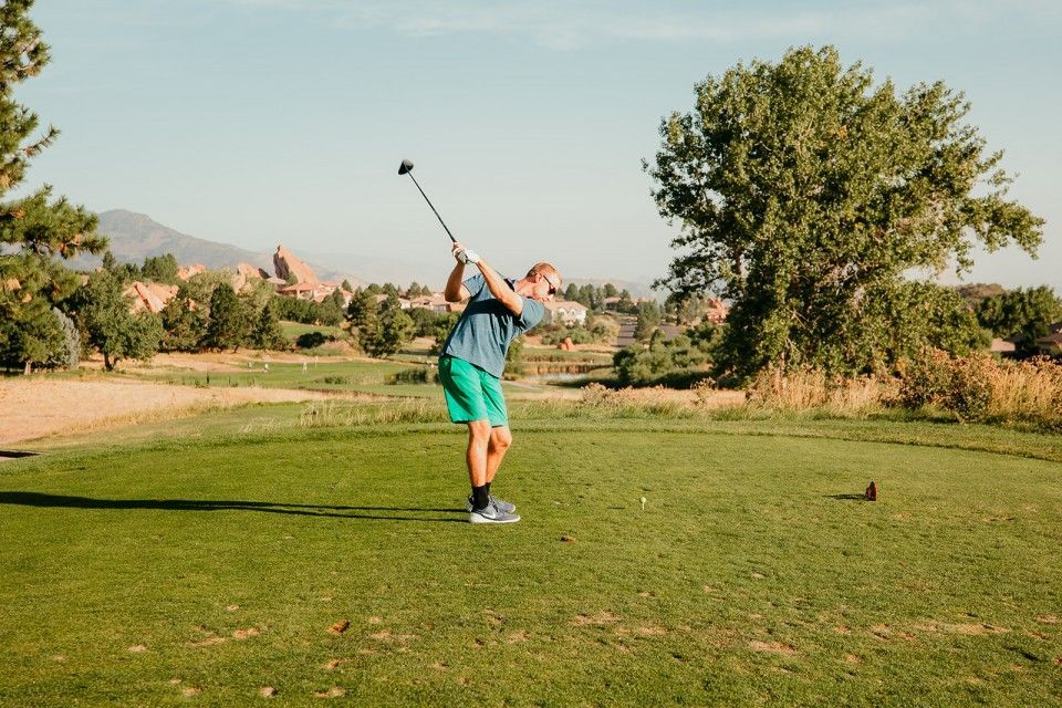 Golfer swinging driver on a golf course, green shorts, tree-lined fairway, sunny day.
