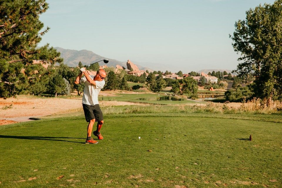 Man swinging a golf club on a green course with mountains in the background. Sunny day.