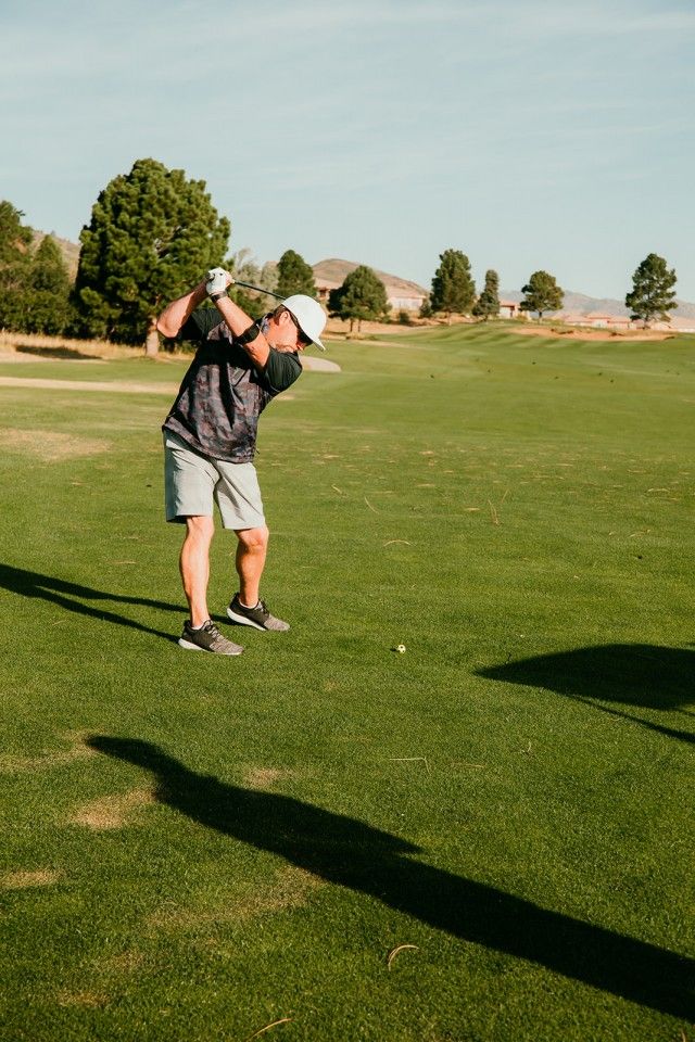 Man swings golf club on a green course, shadow cast on the grass.