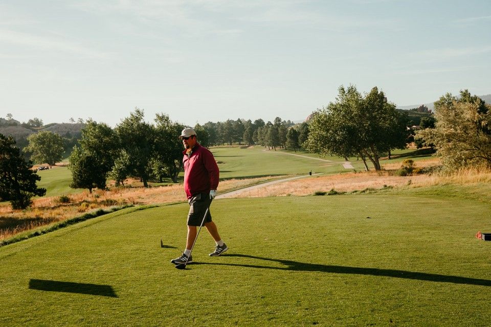 Man on golf course tee box, walks toward ball, wearing red jacket, shorts, and cap on sunny day.
