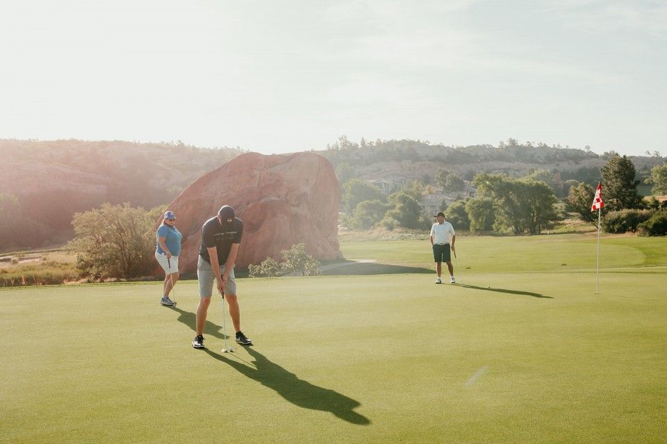 Man putting golf ball on green with two others watching near a flag on a sunny day.