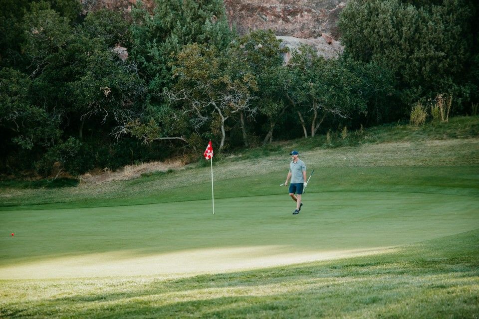 Person walking on green golf course toward the red flag, trees in the background.