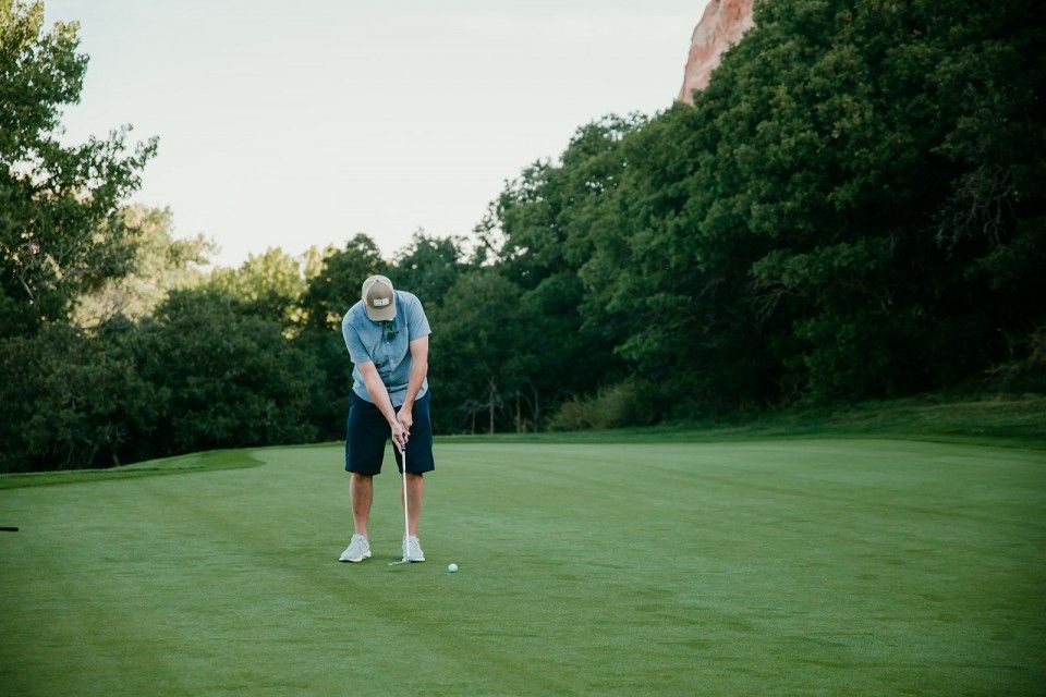 Person putting golf ball on a green with trees and a red rock formation in the background.