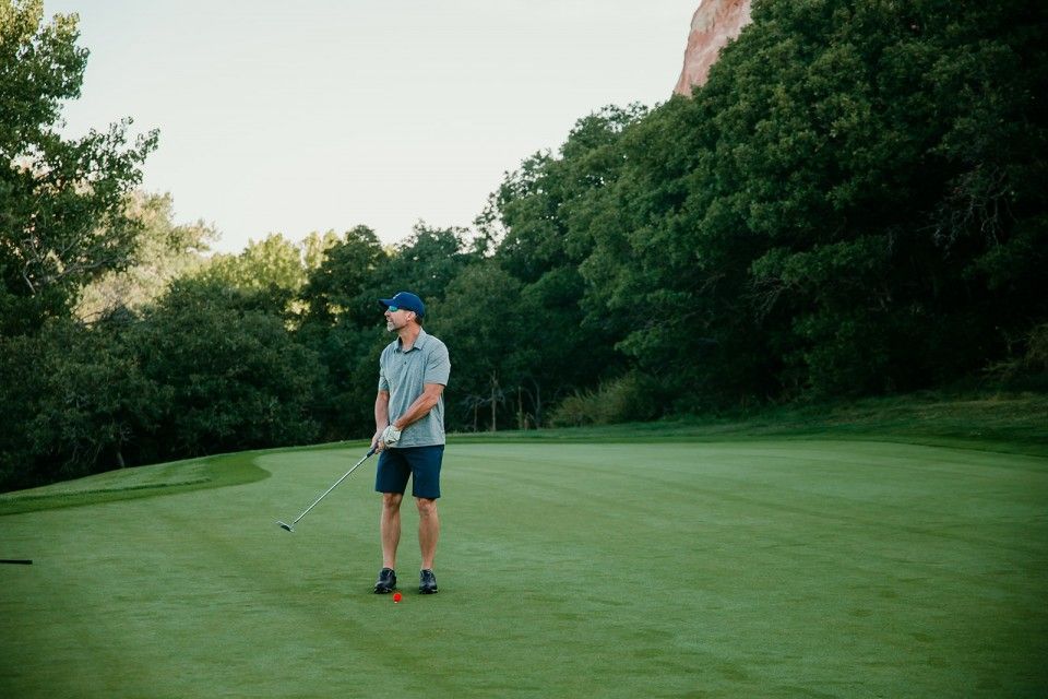 Man on golf course preparing to swing club; green grass, trees, and rock formations in the background.