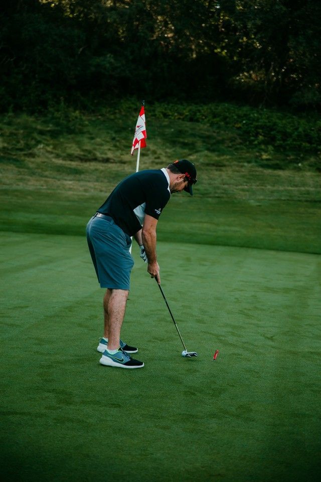 Golfer putting on green, flag in background. Wearing black shirt, blue shorts, dark shoes, and visor.