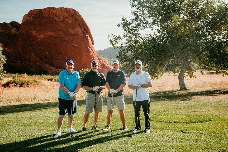 Four men on a golf course pose with clubs; red rock formation in the background.